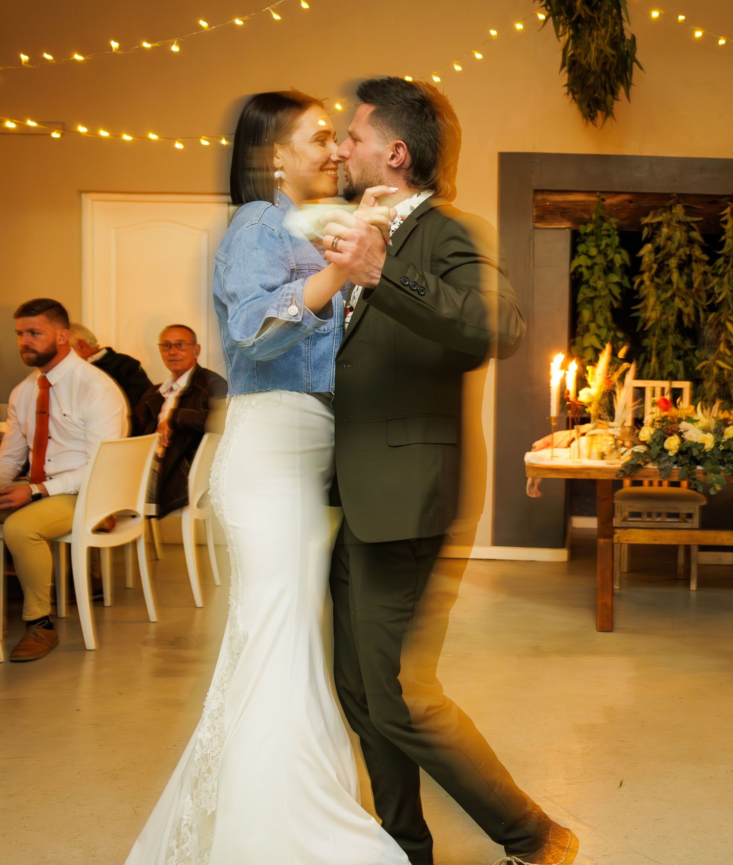 Bride and groom share a romantic first dance in softly lit wedding venue with seated guests.