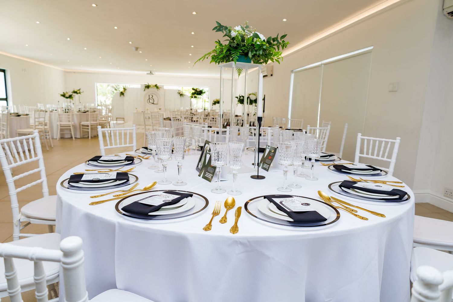 Elegant wedding reception hall with round tables, white linens, gold cutlery, and tall floral centerpieces.