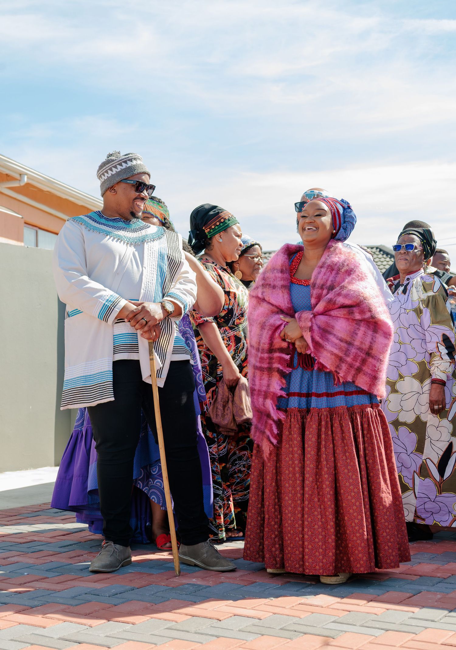 Couple in traditional Xhosa attire with walking stick stands among colorfully dressed guests outdoors.