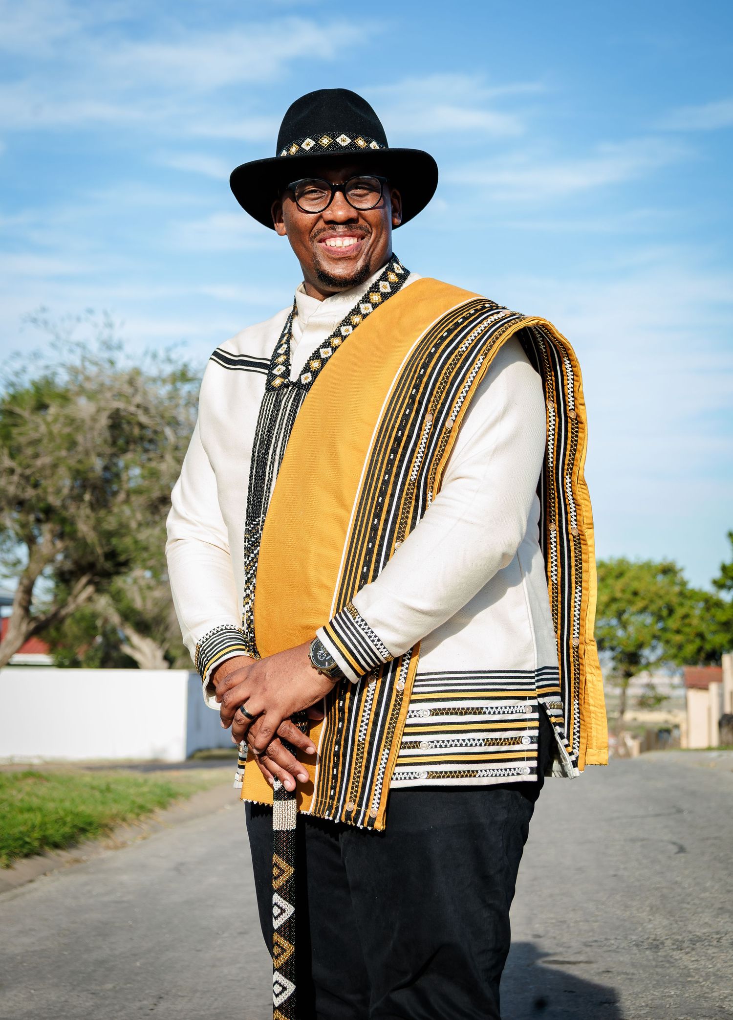 Man smiling confidently in traditional Xhosa white and gold patterned outfit with black wide-brim hat.