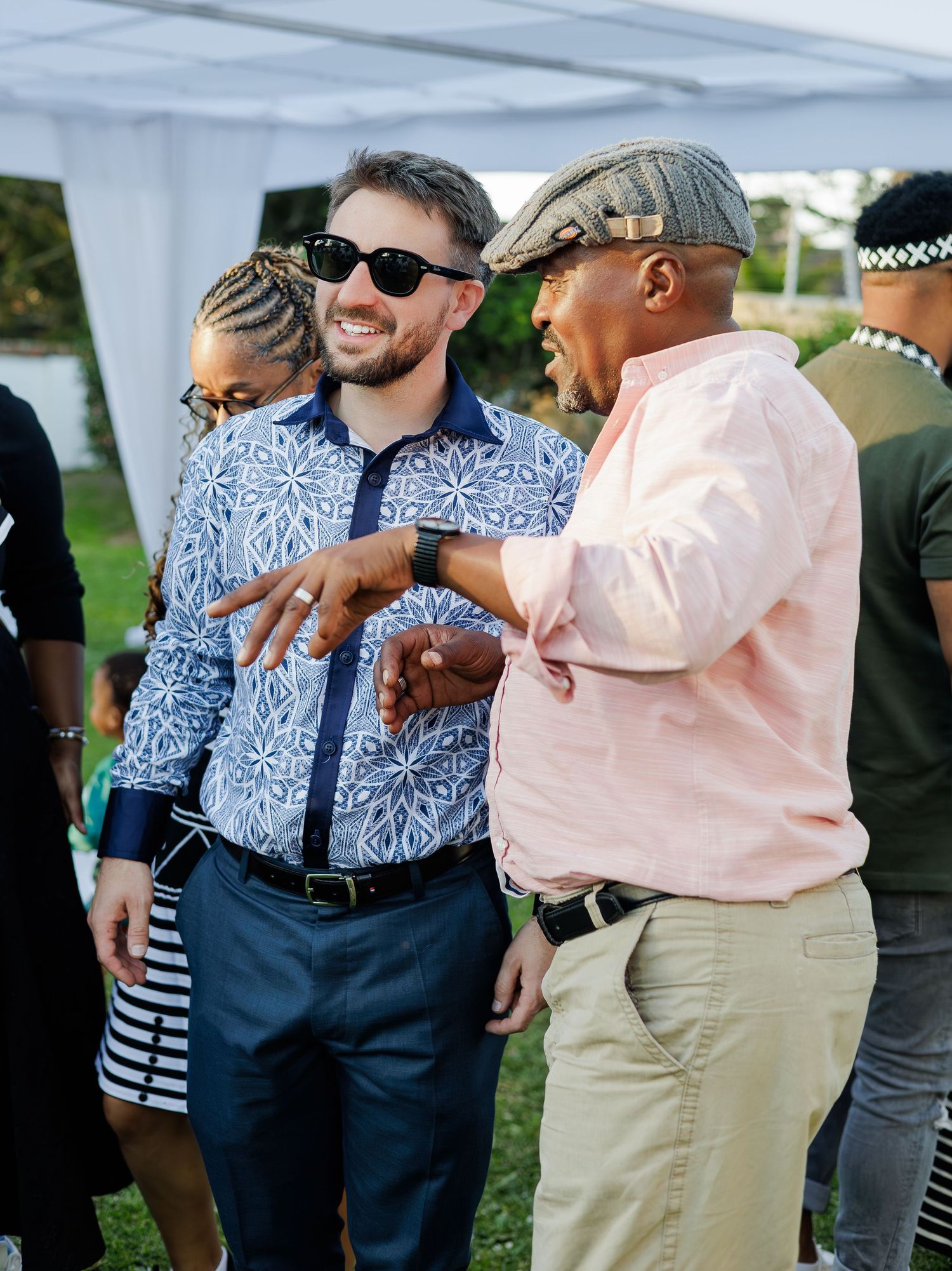 Two men smiling and conversing at an outdoor African cultural celebration wearing patterned shirts.