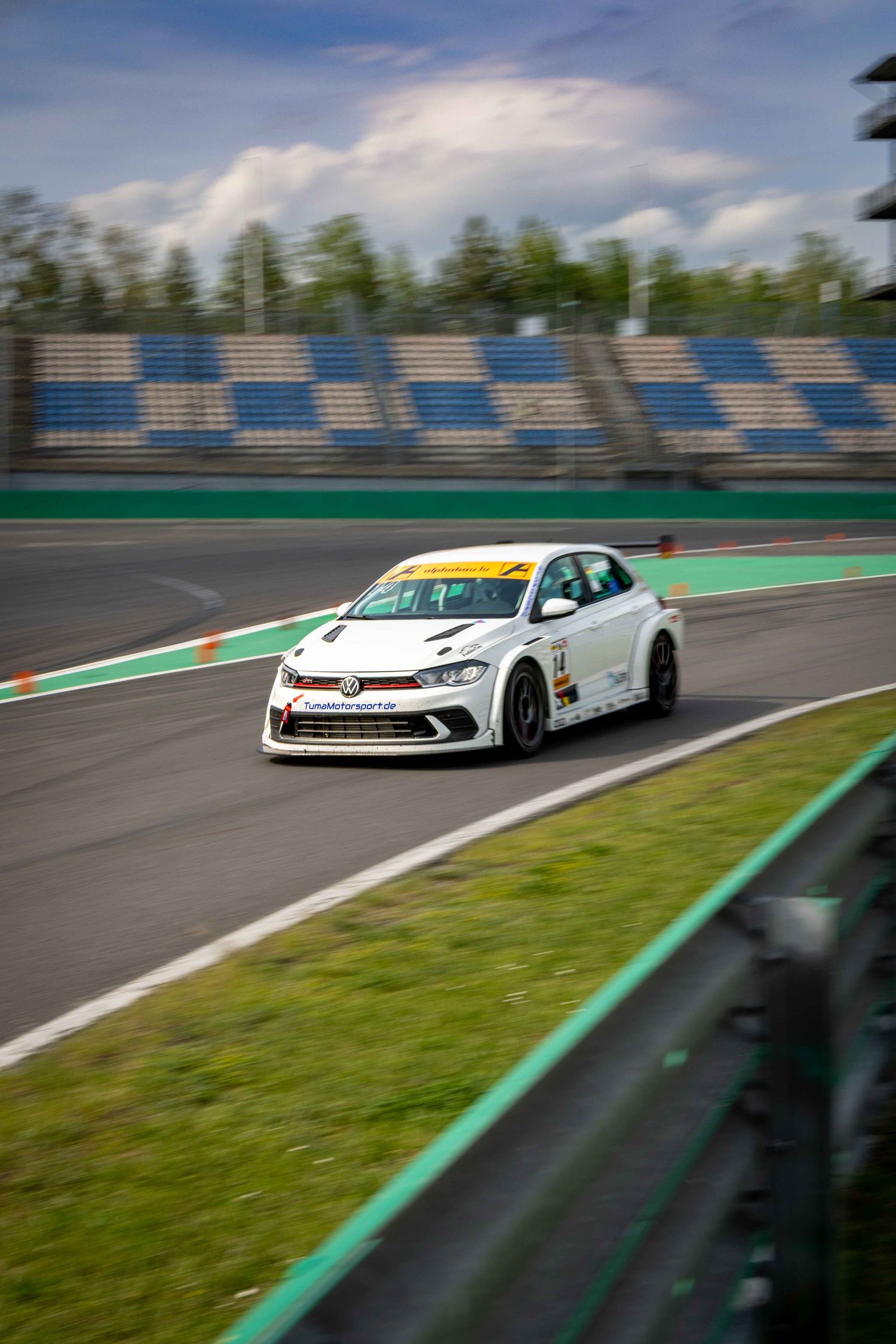 White Volkswagen TCR race car speeding on a circuit track with empty grandstands and cloudy sky backdrop.