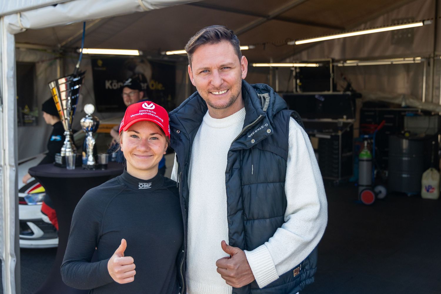 Two people giving thumbs up near racing trophies in a motorsport paddock garage area.