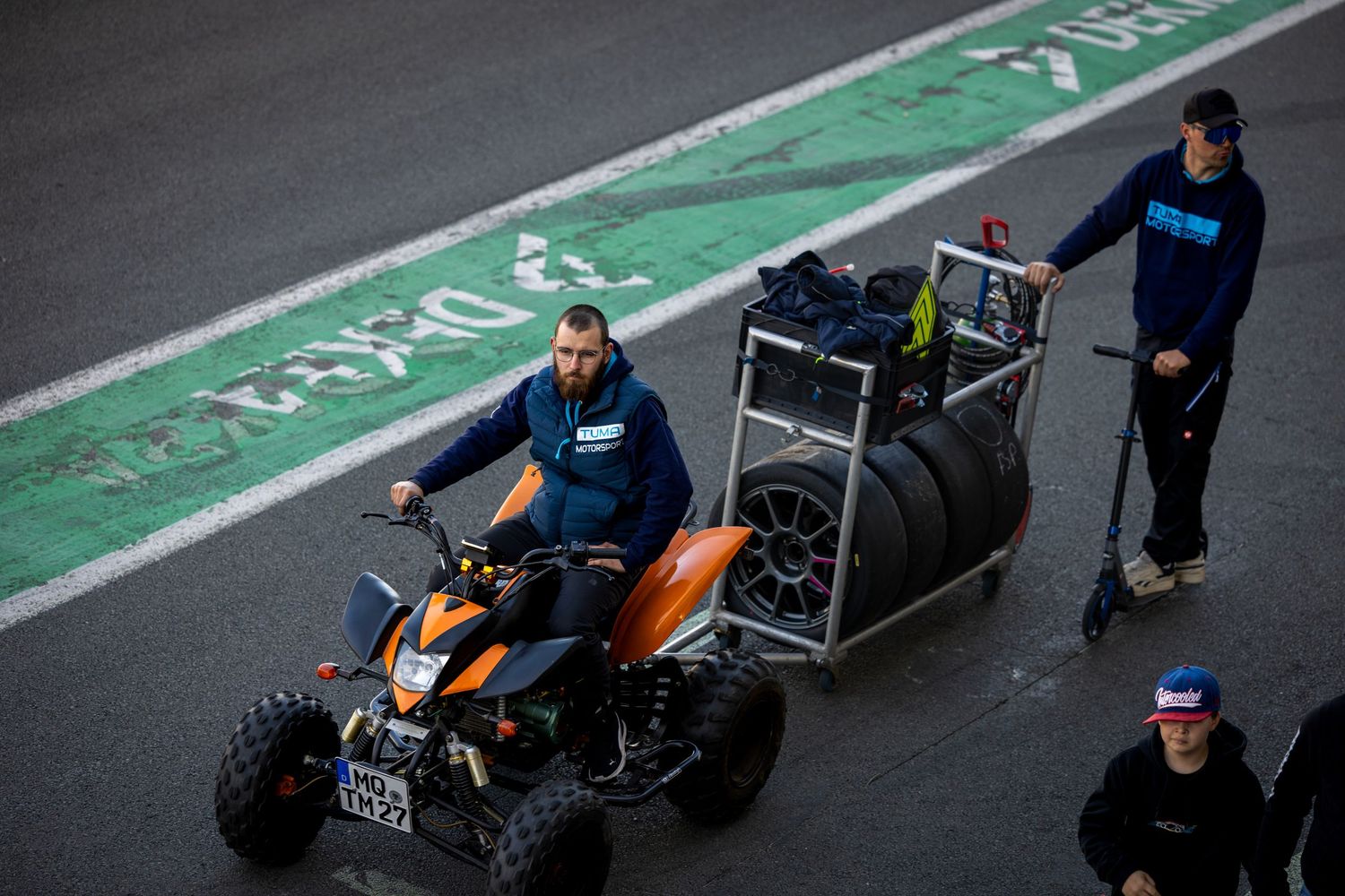 Racing team crew member rides orange ATV quad bike hauling tires in pit lane during motorsport event.