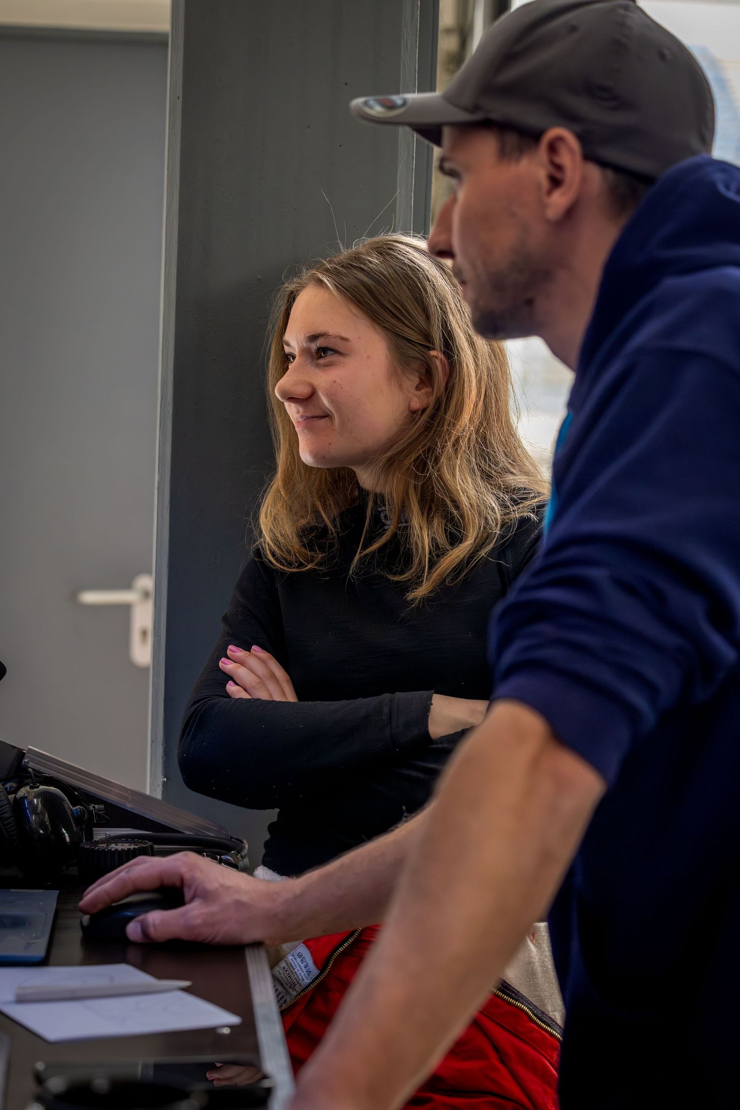 A smiling woman with crossed arms stands next to a man in a blue hoodie and cap working at a desk indoors.