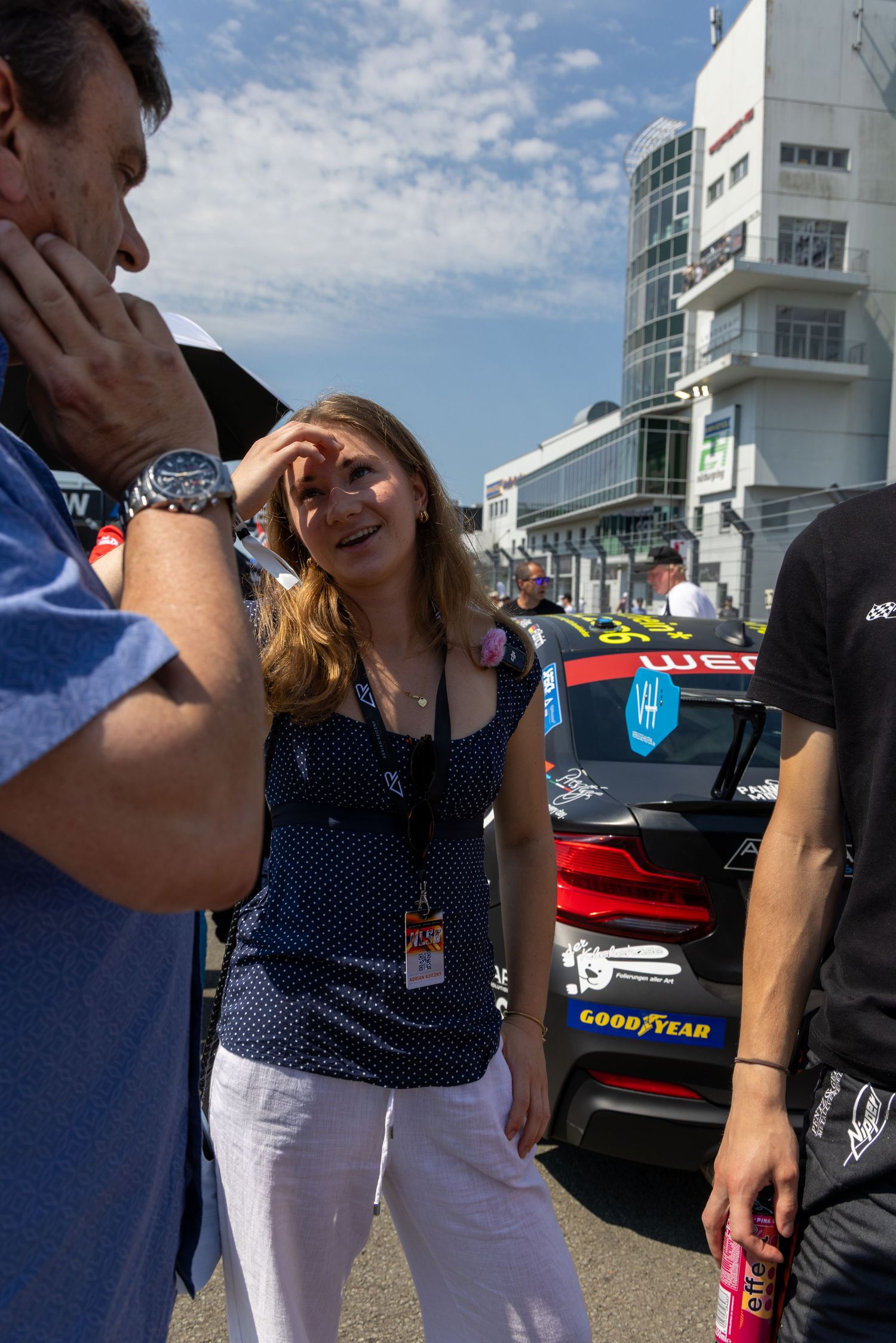 Woman with lanyard smiling at a racing event, with a Goodyear-sponsored race car visible in the background.