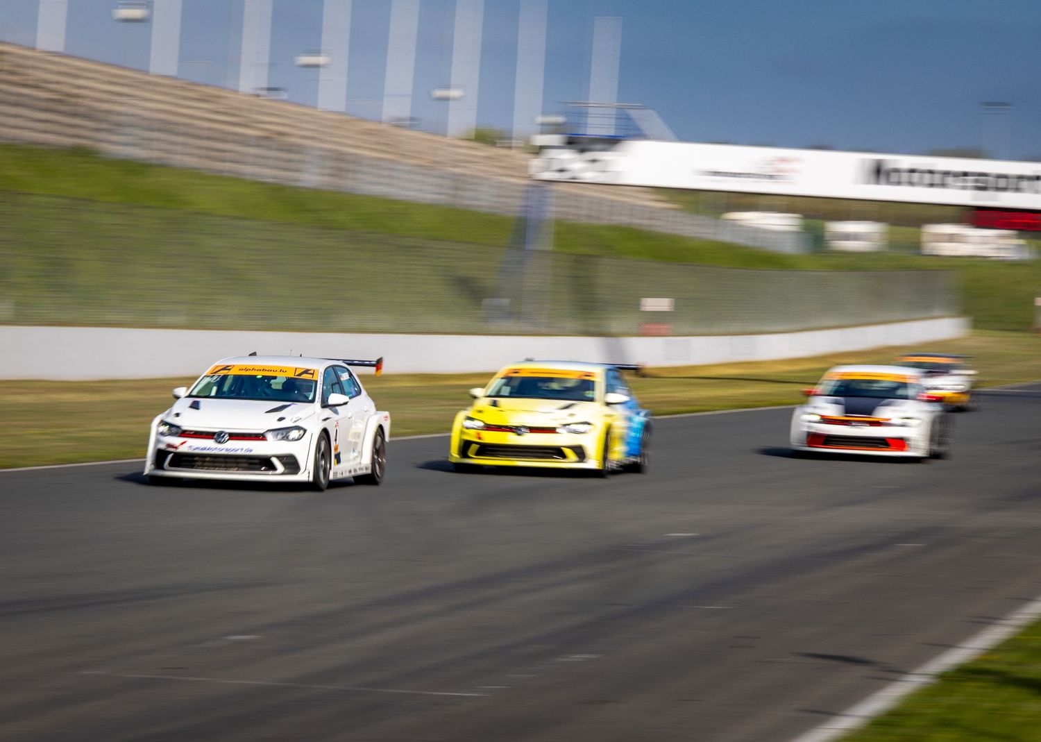 Racing cars compete on a track, led by a white VW Golf TCR followed by yellow and blue competitors.