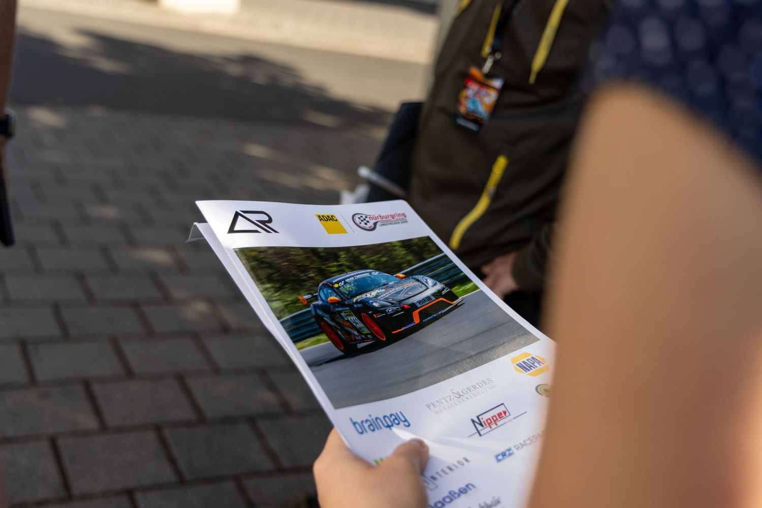 Person holding a racing event program featuring a sports car on a track at an outdoor motorsport venue.