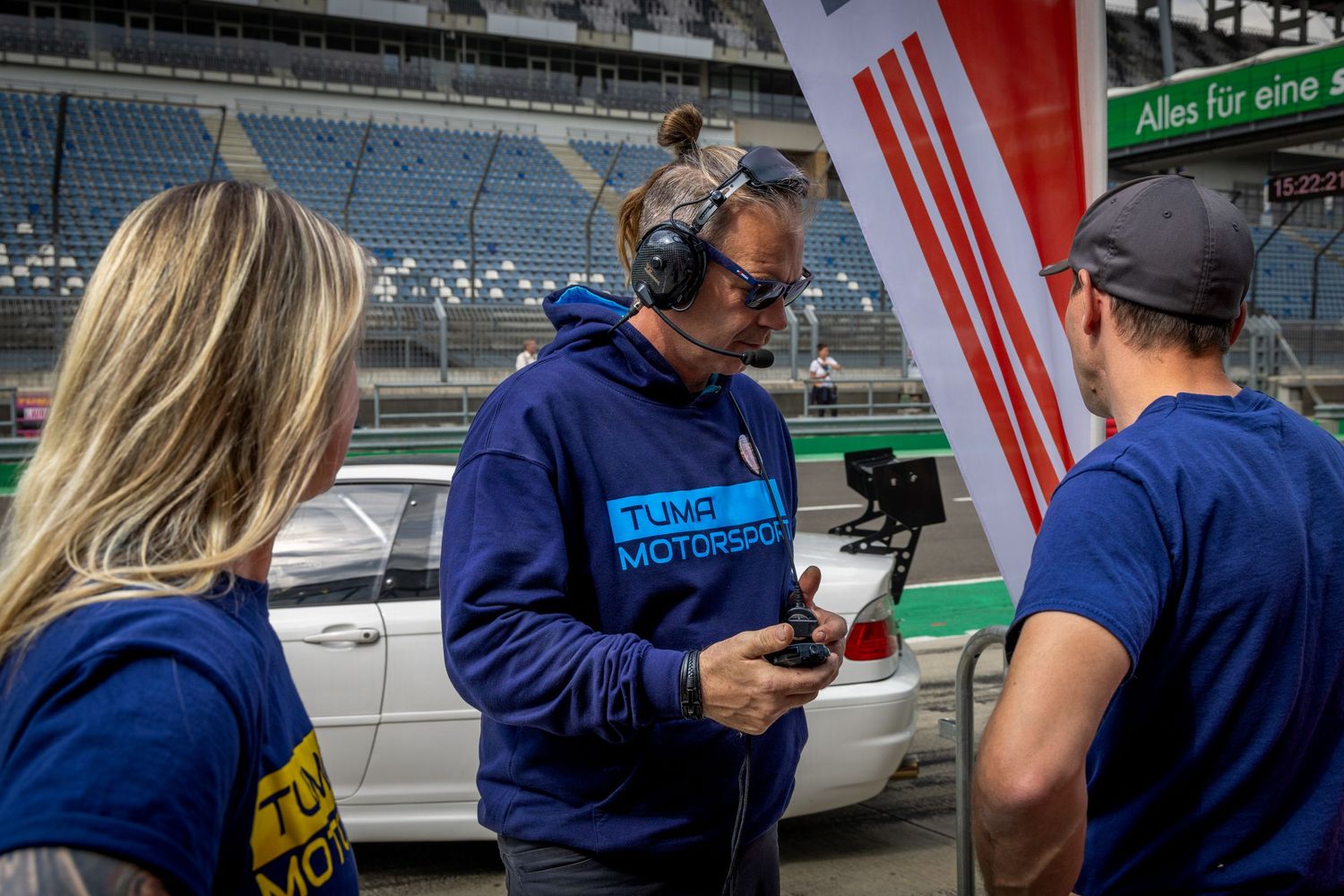 Three motorsport crew members in blue hoodies reviewing footage on a camera at a racing circuit pit lane.