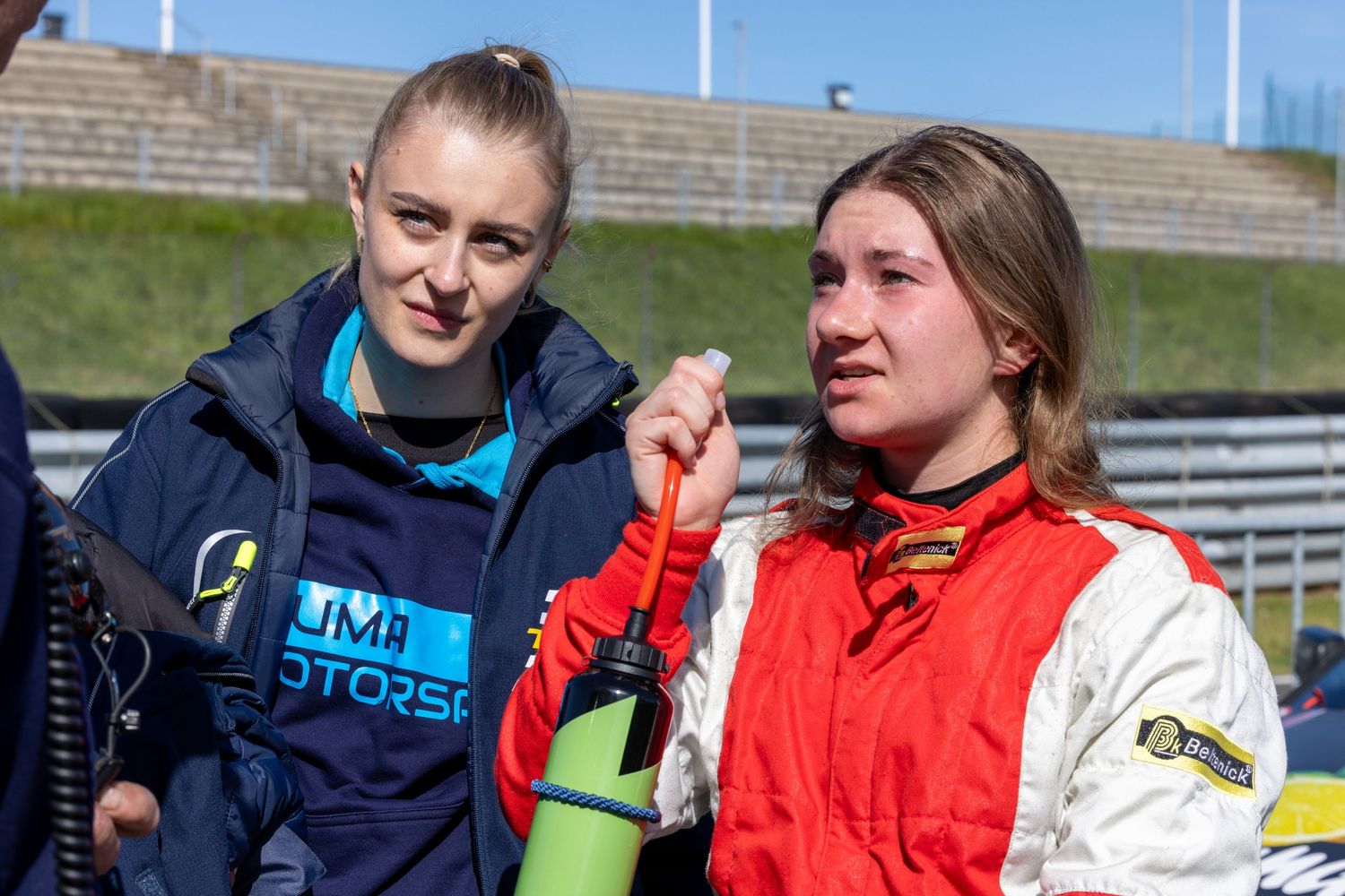 Two young female racing drivers in motorsport attire discuss race strategy at a circuit event.