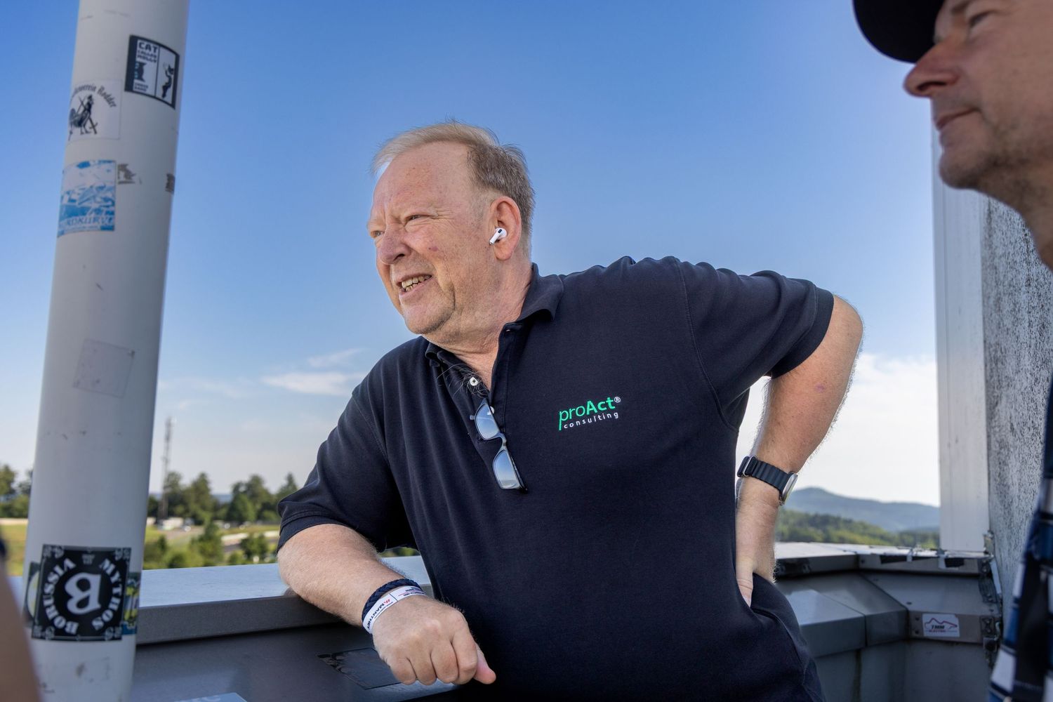 Man in dark polo shirt smiling outdoors near a pole on a sunny day with scenic hills in background.