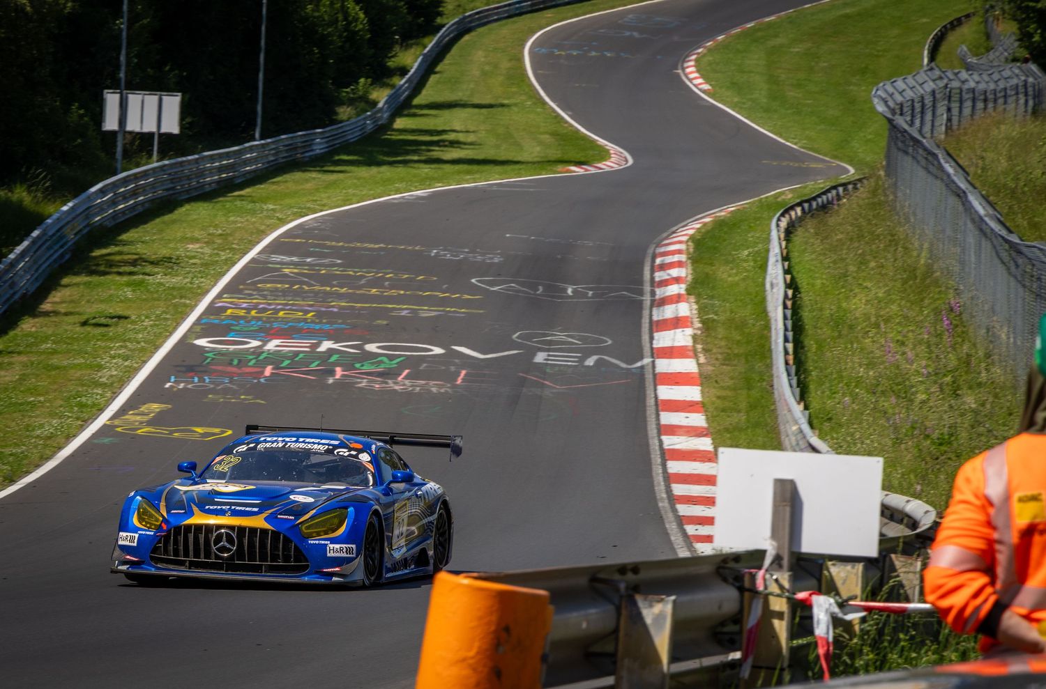 Blue Mercedes-AMG GT3 racing car competing on the iconic Nurburgring Nordschleife circuit on a sunny day.