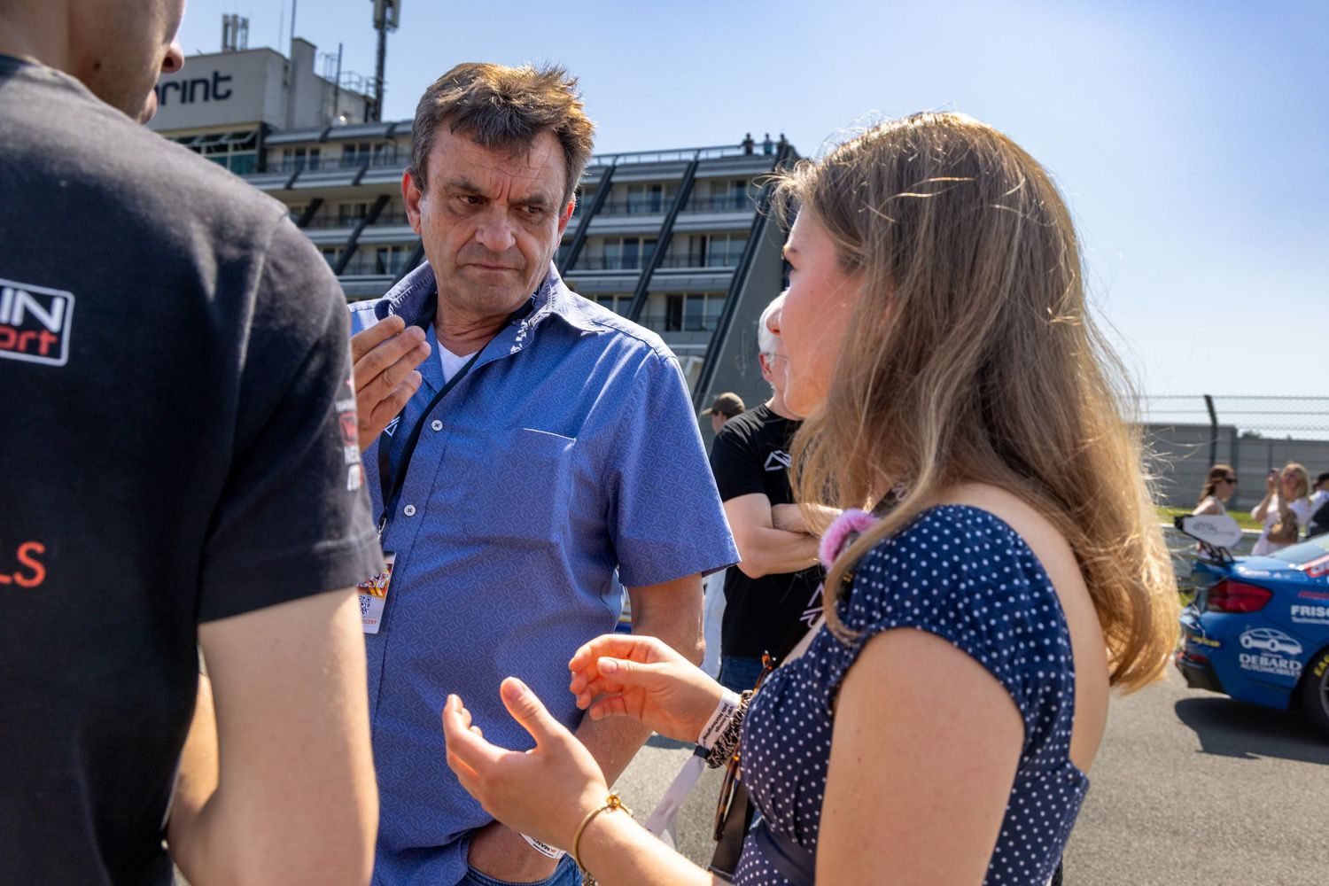 Three people engaged in conversation outdoors at a sunny racing circuit event, with a blue car visible in the background.