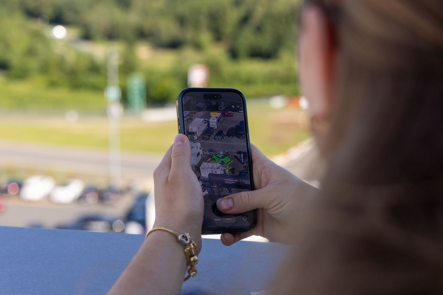 Woman holding smartphone displaying game app outdoors, with green trees and parking lot visible in background.