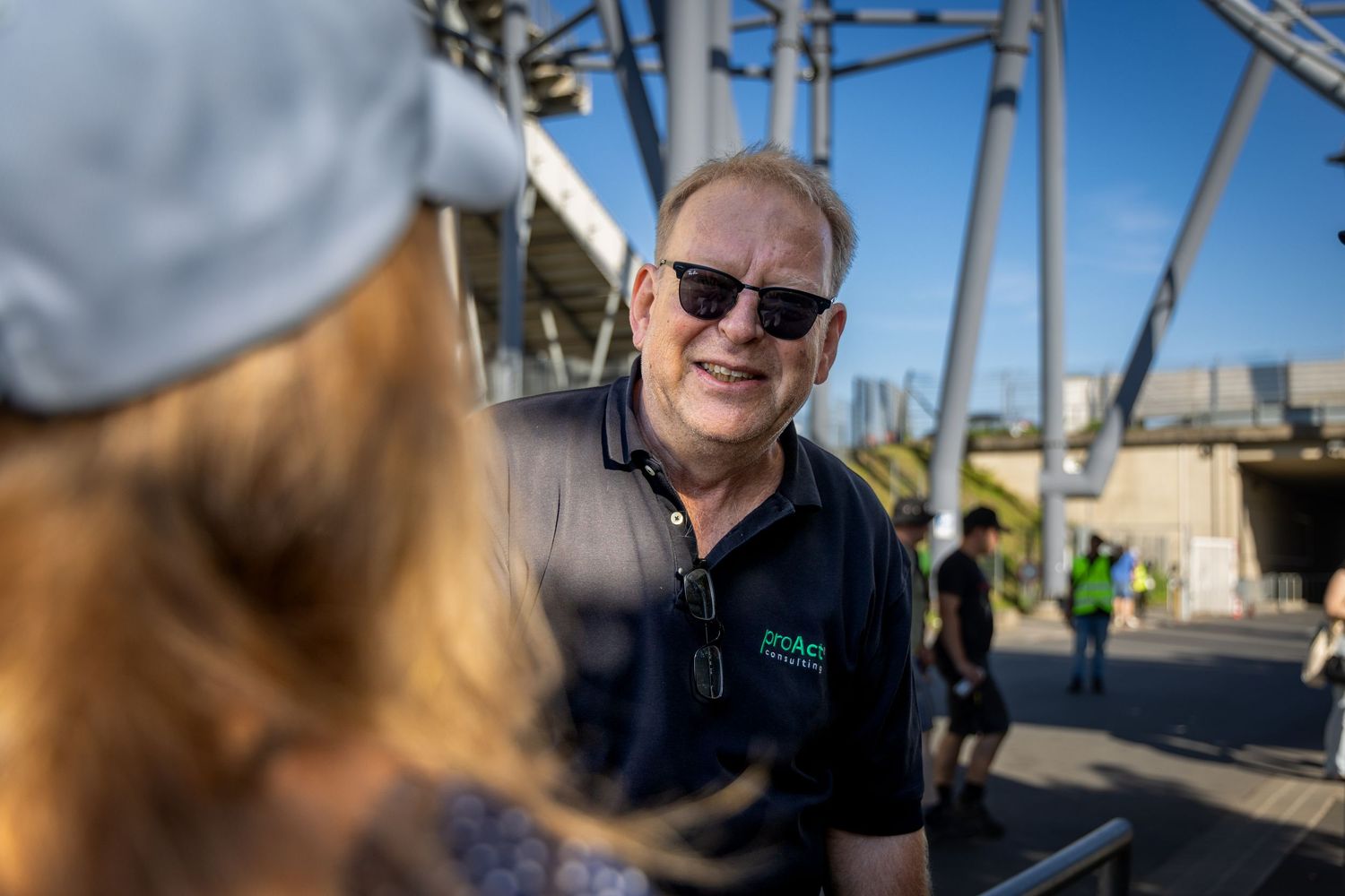 A smiling man wearing sunglasses and a dark polo shirt converses outdoors near a modern steel structure on a sunny day.