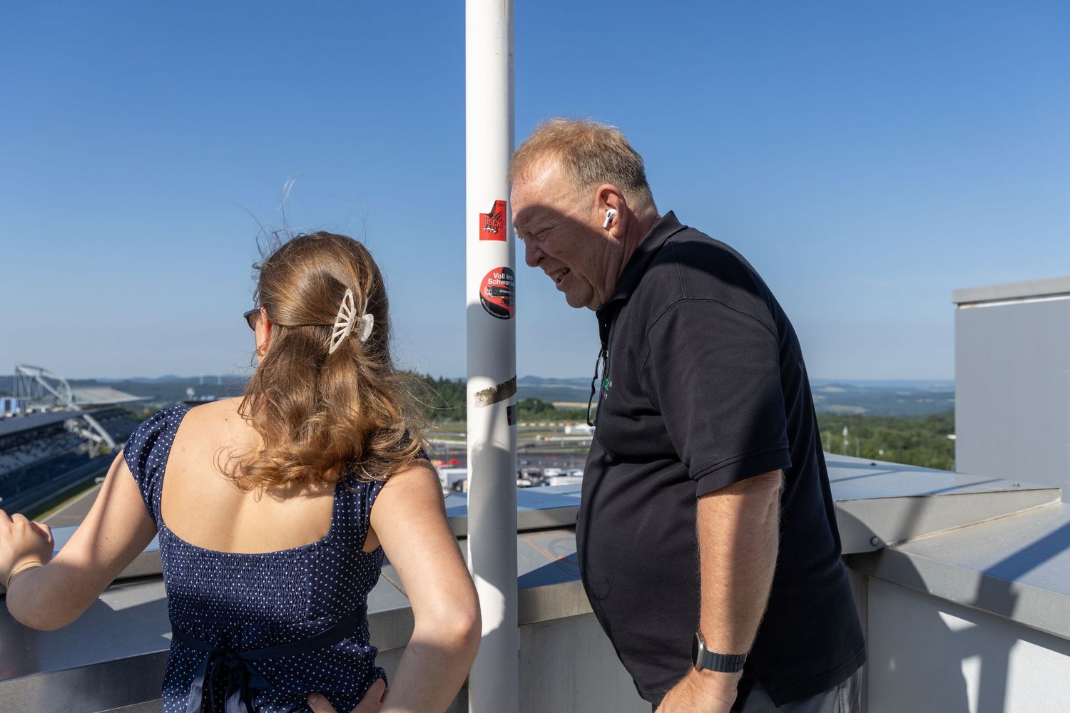 Two people stand on an outdoor observation deck on a sunny day, with a scenic landscape visible in the background.