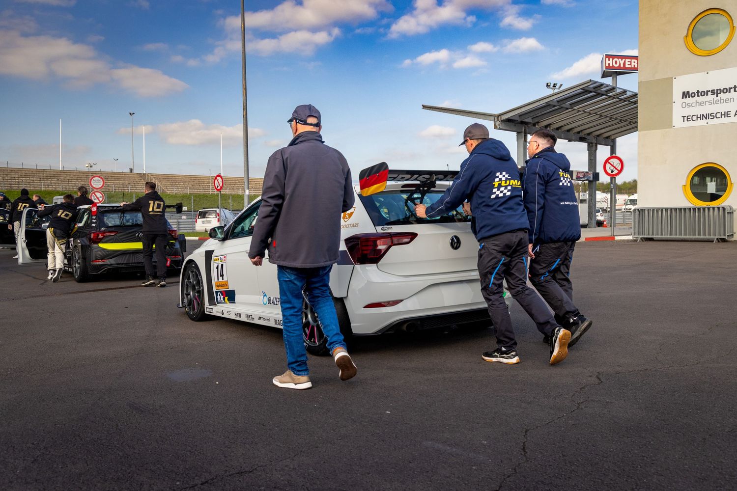 Mechanics push a German-flagged race car in a motorsport pit lane during a racing event.