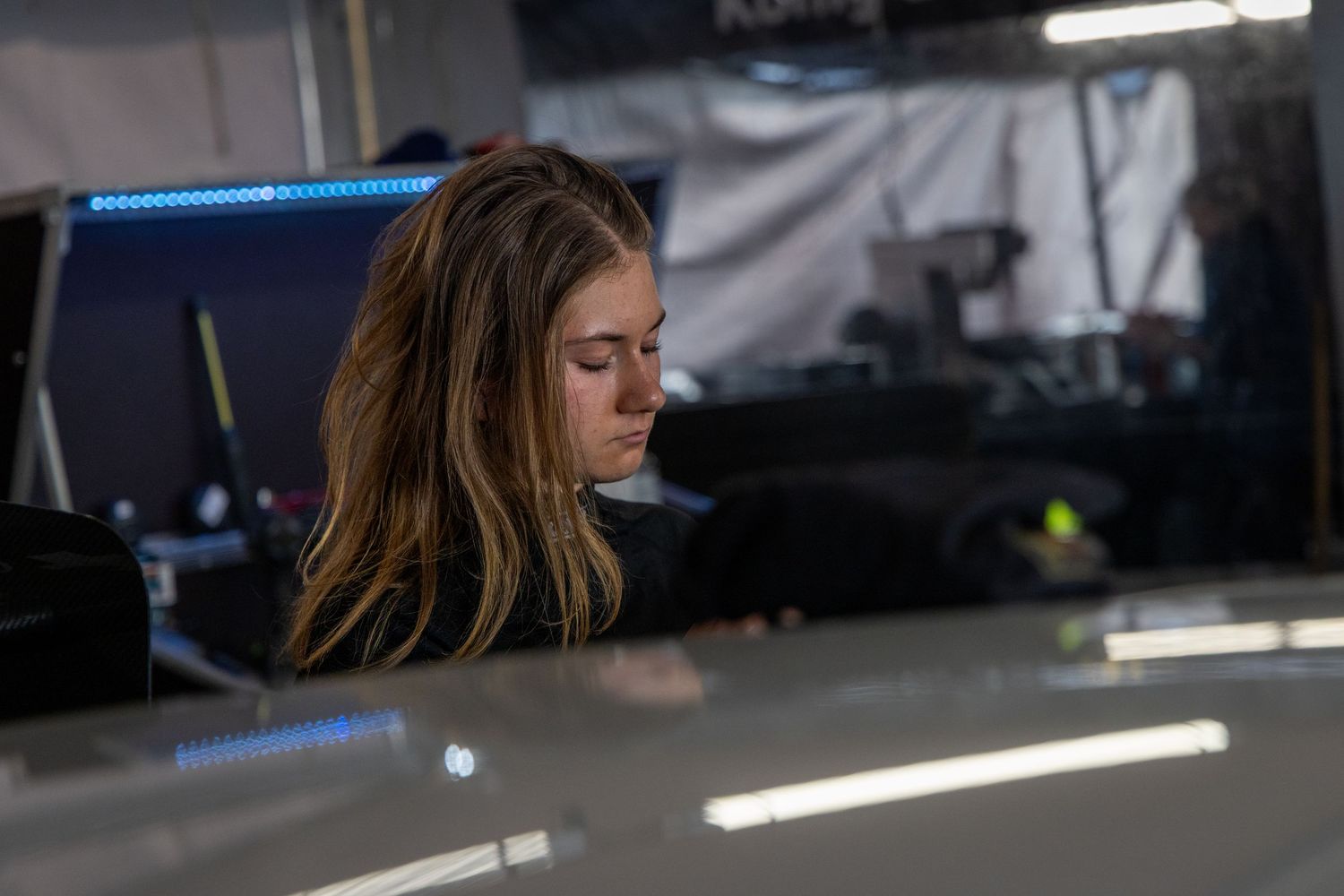 Young woman with long brown hair working on a shiny car in a dimly lit auto shop.
