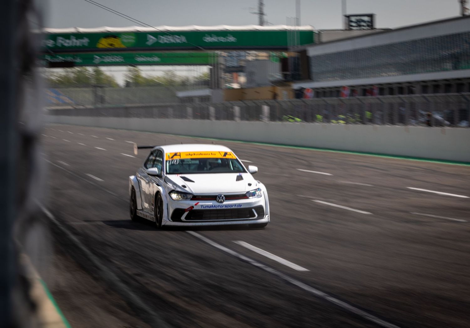 A white racing car speeds along a track at a motorsport circuit with green barriers and grandstands in the background.