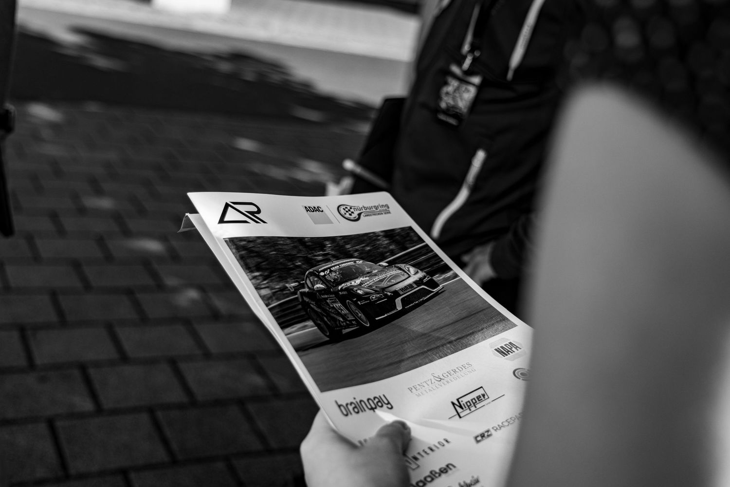 Person holding a motorsport event program booklet outdoors, black and white photography at a racing venue.