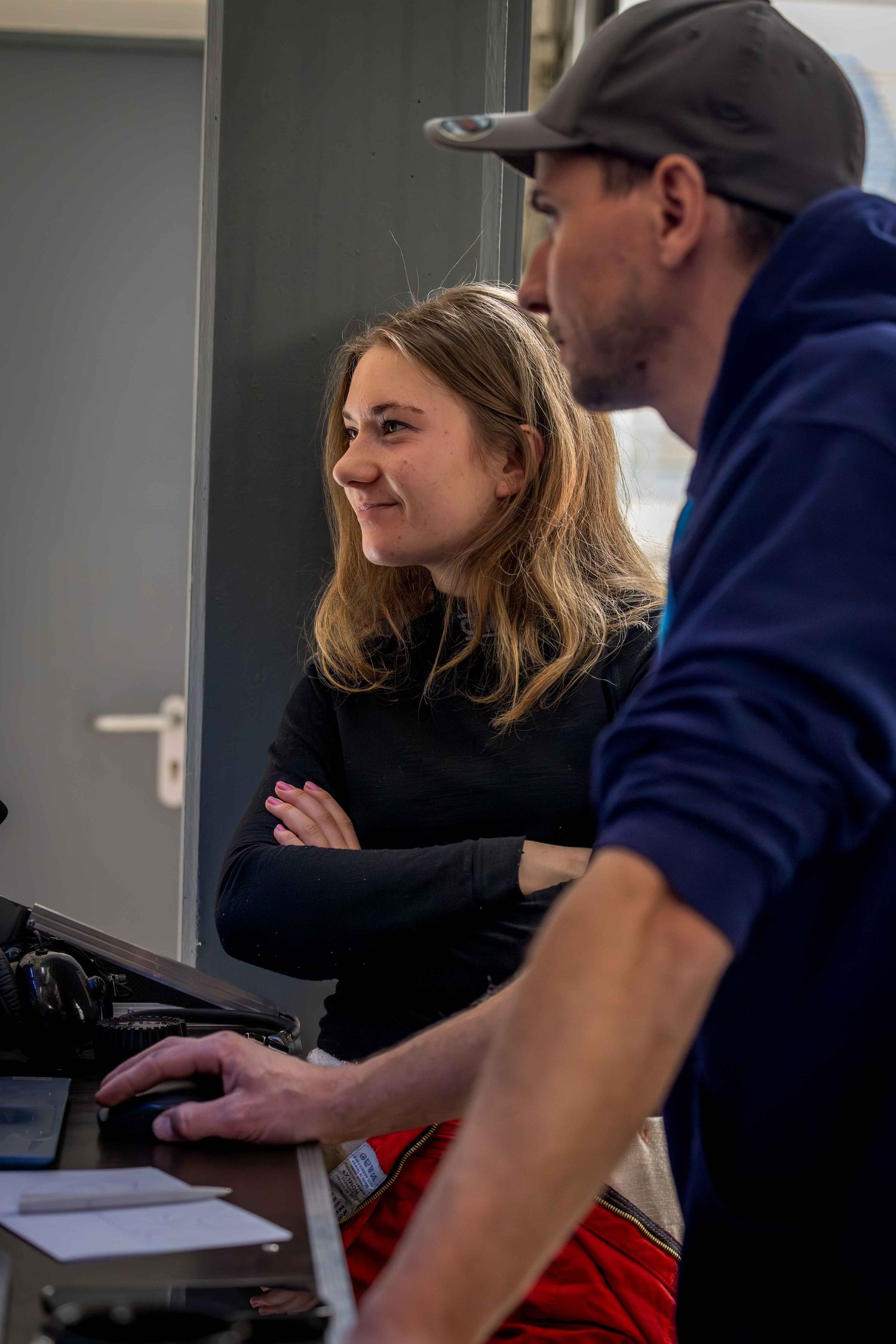 A smiling woman with crossed arms stands next to a man in a cap working at a desk in an office setting.