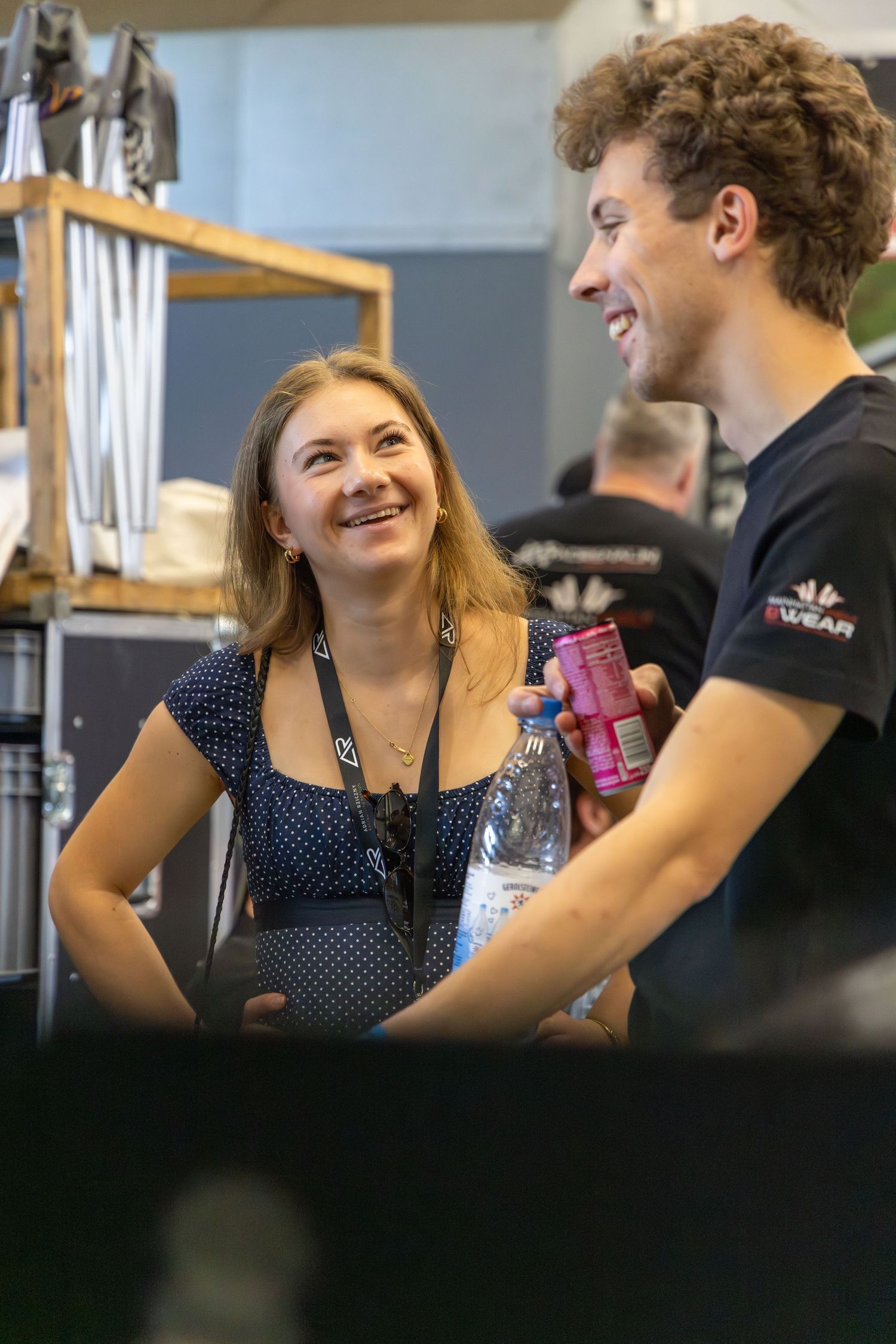 A smiling woman with a lanyard chats with a curly-haired man holding a drink at an indoor event.