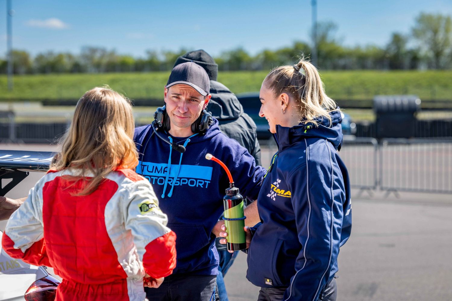 Three people in motorsport jackets chat at a racing event on a sunny day near a track barrier.