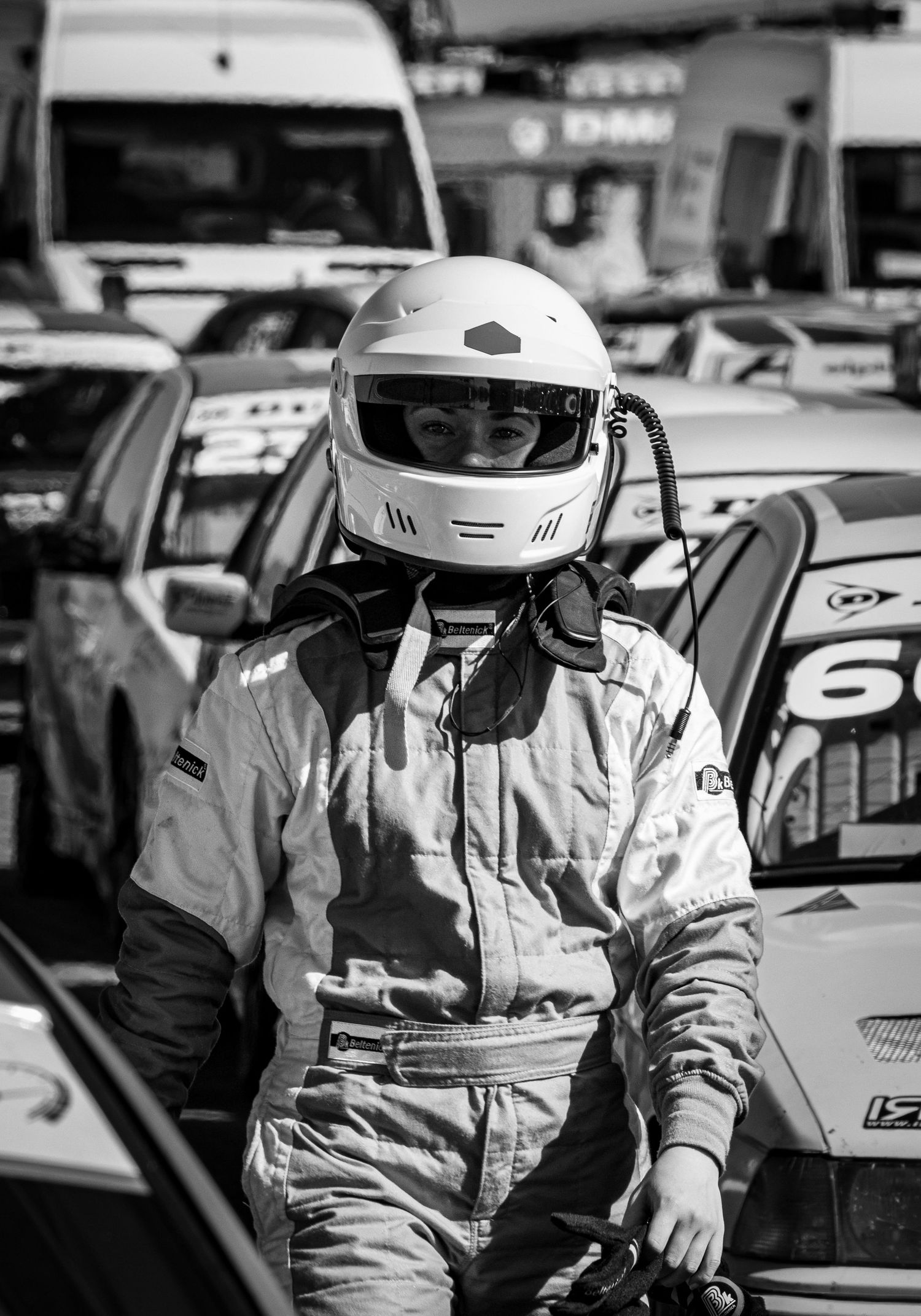 Black and white photo of a race car driver in full gear and white helmet standing near race cars at a motorsport event.