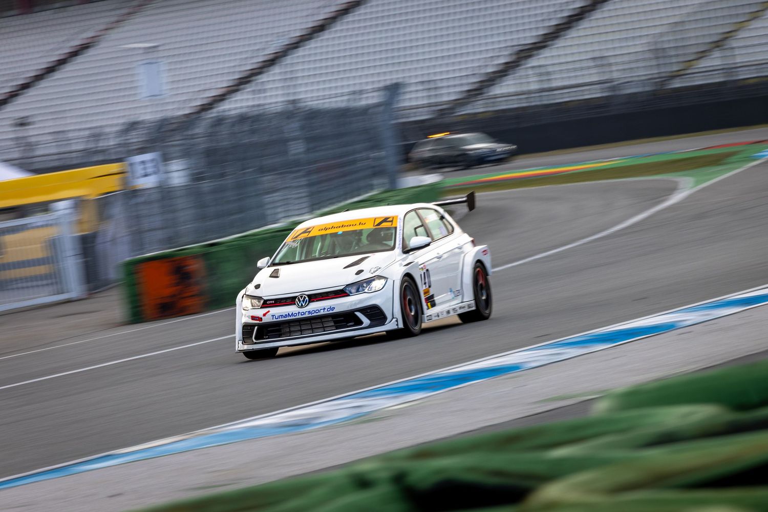 A white Volkswagen race car speeds around a curve on a motorsport track with empty grandstands in the background.