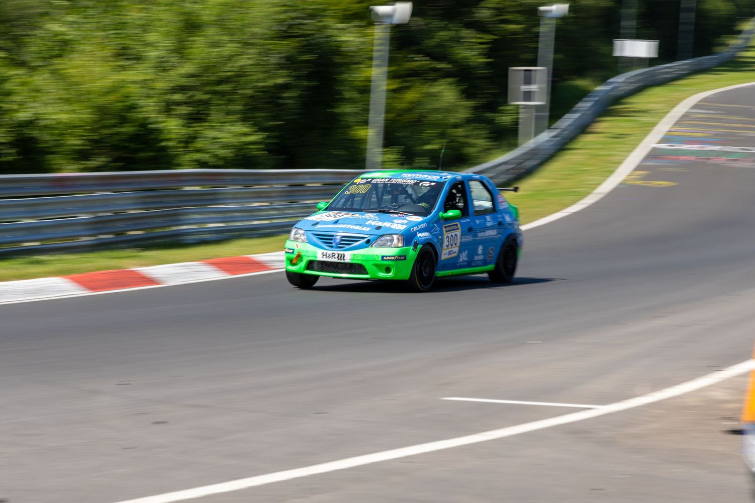 Green and blue race car speeding on Nurburgring circuit track surrounded by lush trees on a sunny day.