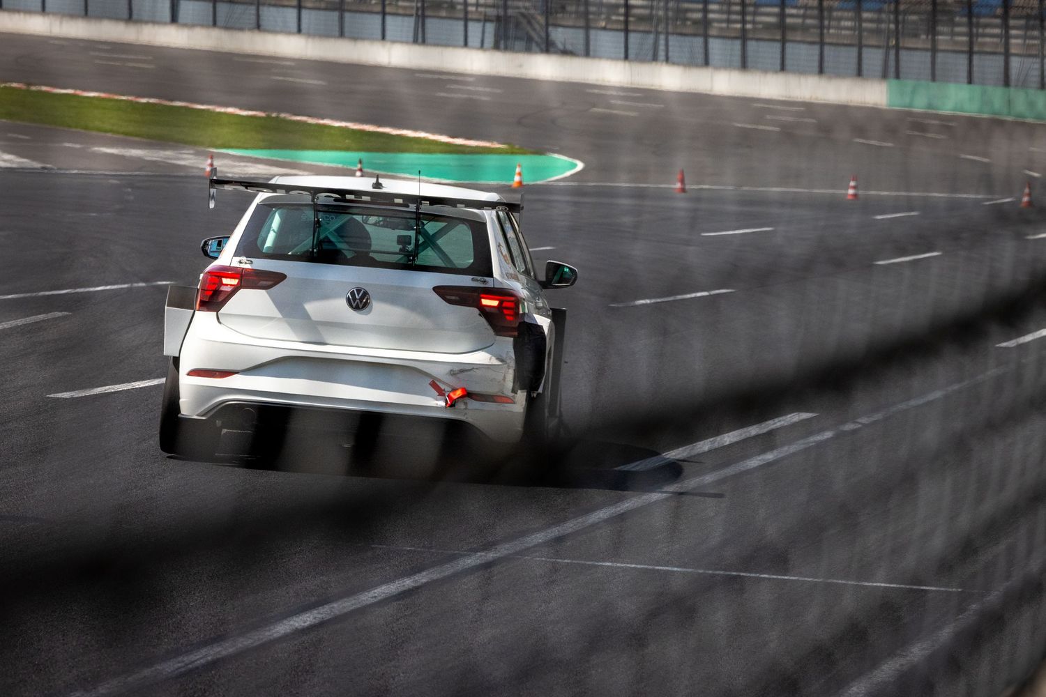 A white VW race car speeds around a wet racing circuit track, viewed from behind, with a rooftop spoiler.