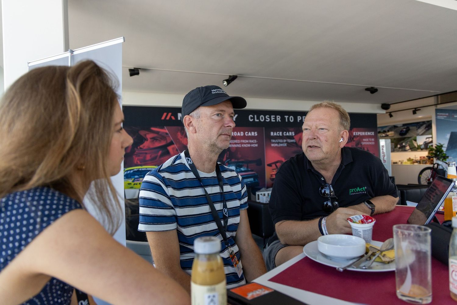 Three people engaged in conversation at an outdoor dining table at a racing event, with promotional banners in the background.