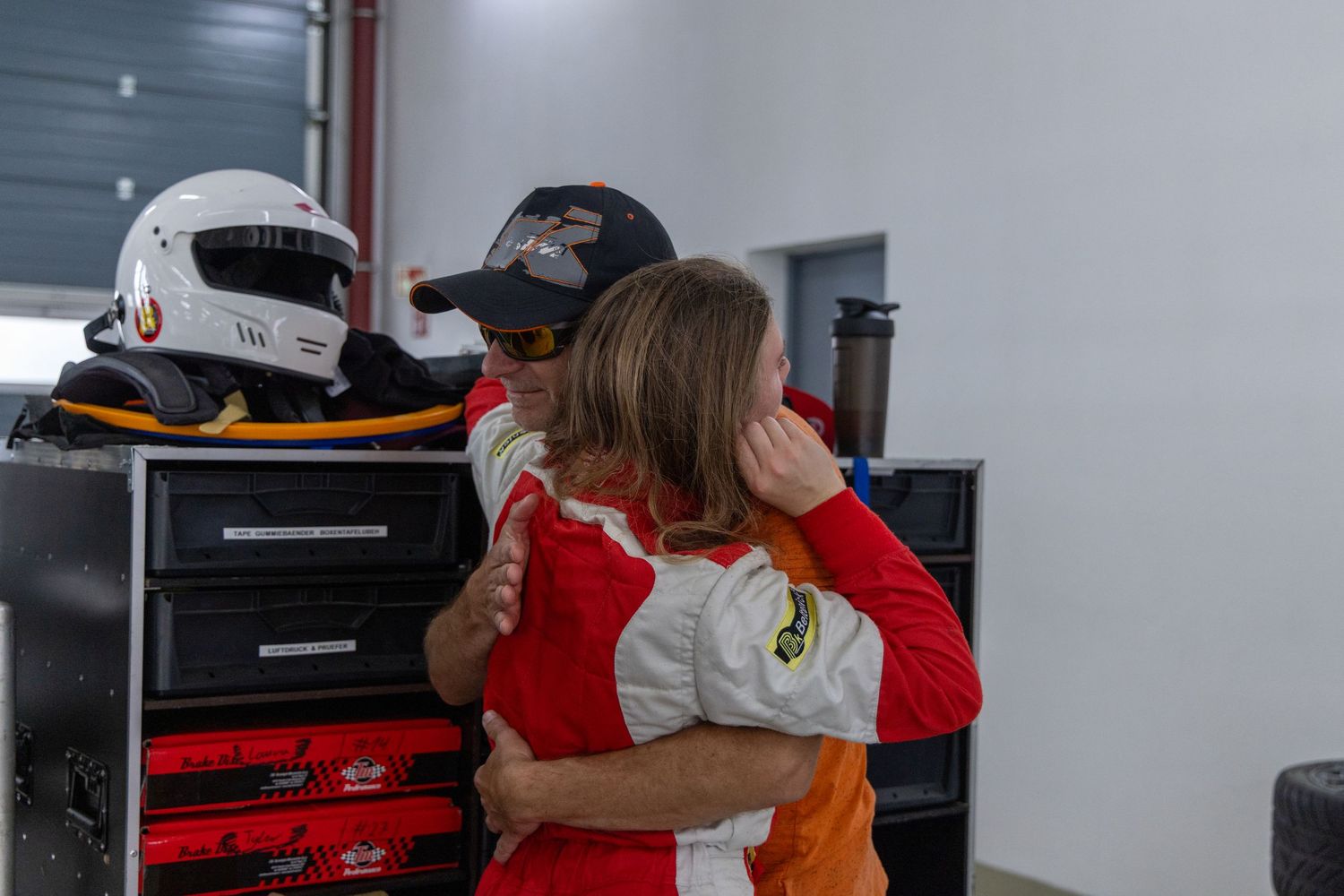 Two racing drivers in colorful suits share a warm embrace in a motorsport garage with helmets visible in the background.