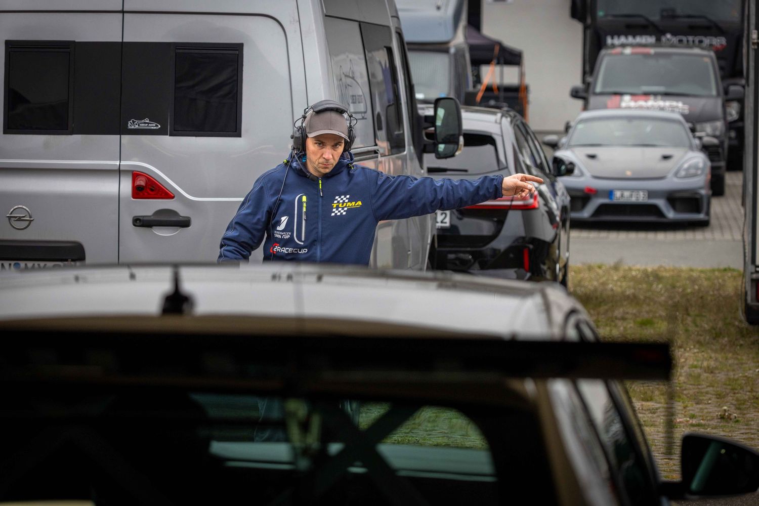 A race marshal in a blue jacket and headset directs traffic in a busy paddock area with vehicles parked nearby.