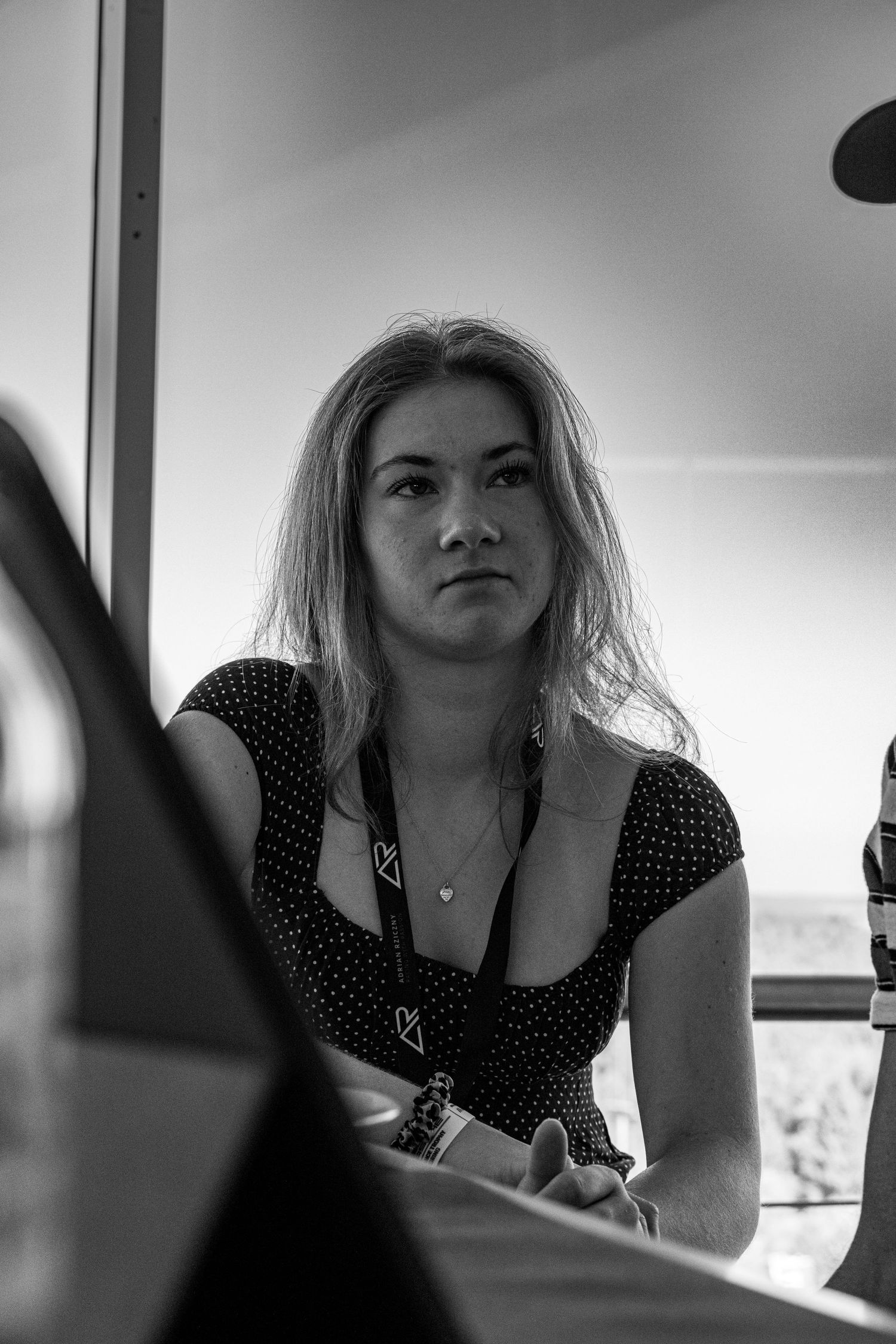 Black and white portrait of a young blonde woman wearing a polka dot dress and lanyard, seated indoors.