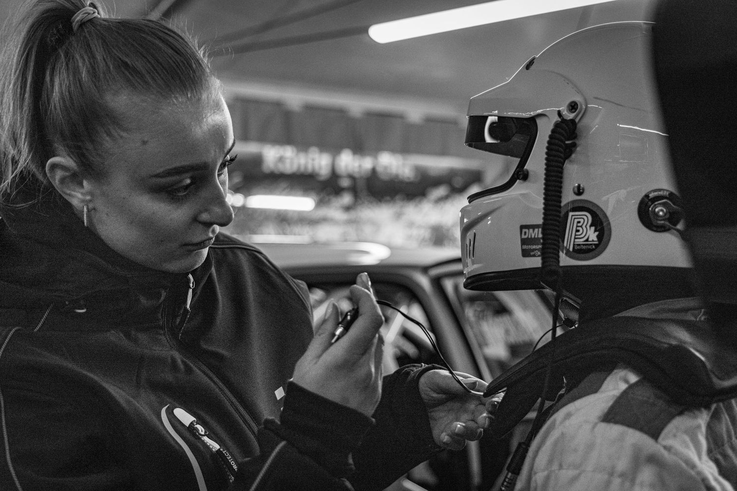 Black and white photo of a woman adjusting a racing helmet on a driver inside a car at a motorsport event.