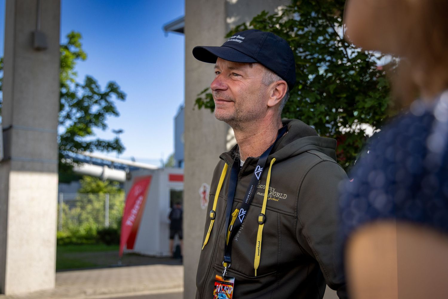 Middle-aged man in dark cap and olive jacket with lanyards stands outdoors near pillars on a sunny day.