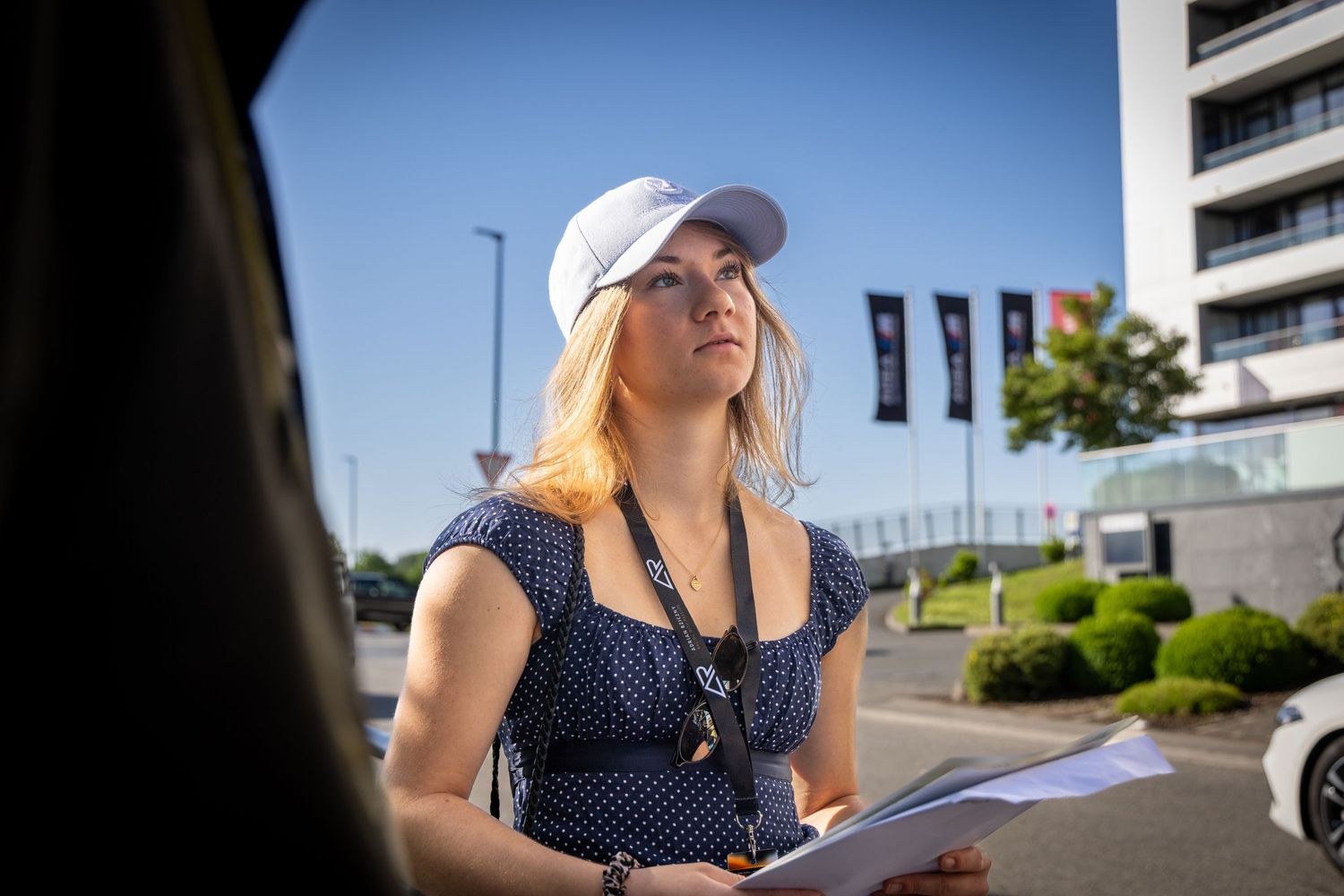 Blonde woman in white cap and polka dot dress holds clipboard outdoors near modern building on sunny day.