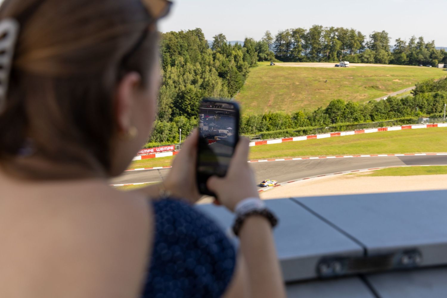 A spectator photographs a motorcycle race on a winding track from the grandstand at a motorsport circuit.