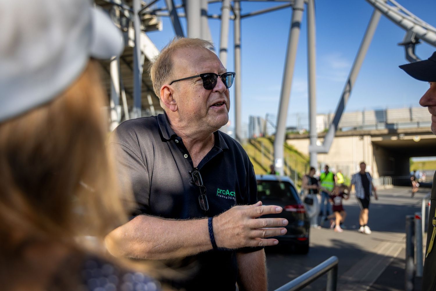Man in black polo shirt with sunglasses holds camera outdoors near a large steel structure on a sunny day.