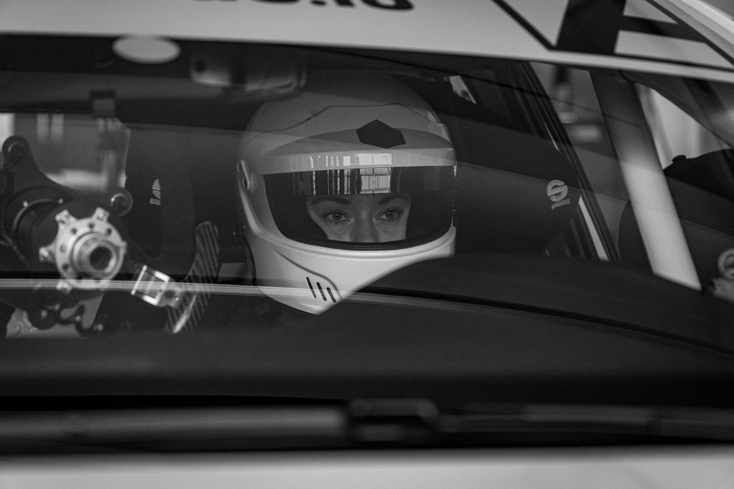 Black and white photo of a race car driver wearing a helmet, seated inside a racing vehicle.