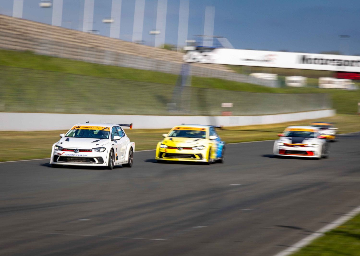 Racing cars compete on a circuit track, led by a white VW Golf TCR followed by yellow and blue vehicles.