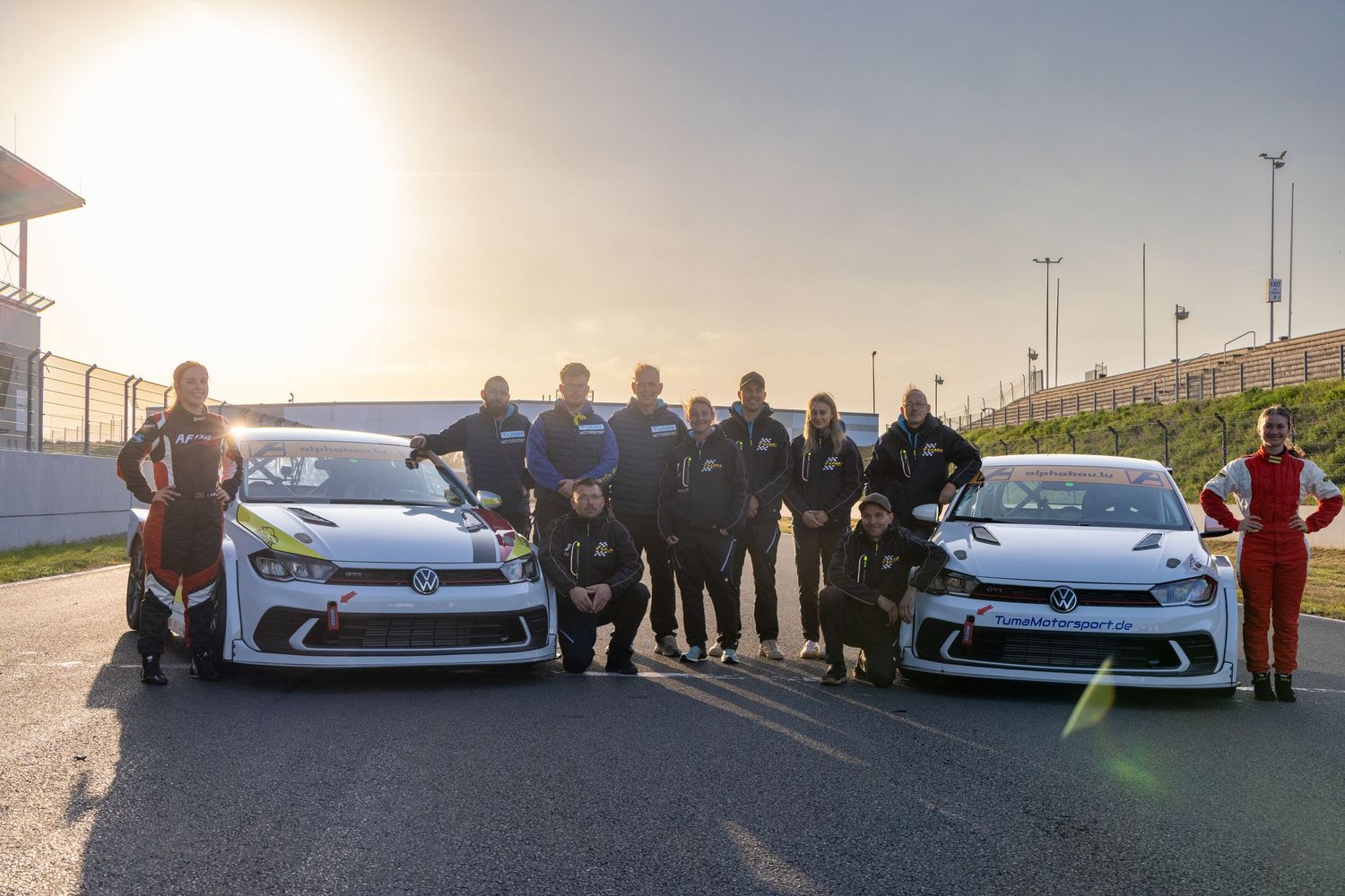 Racing team posing with two VW Polo GTI race cars on a track at sunset.