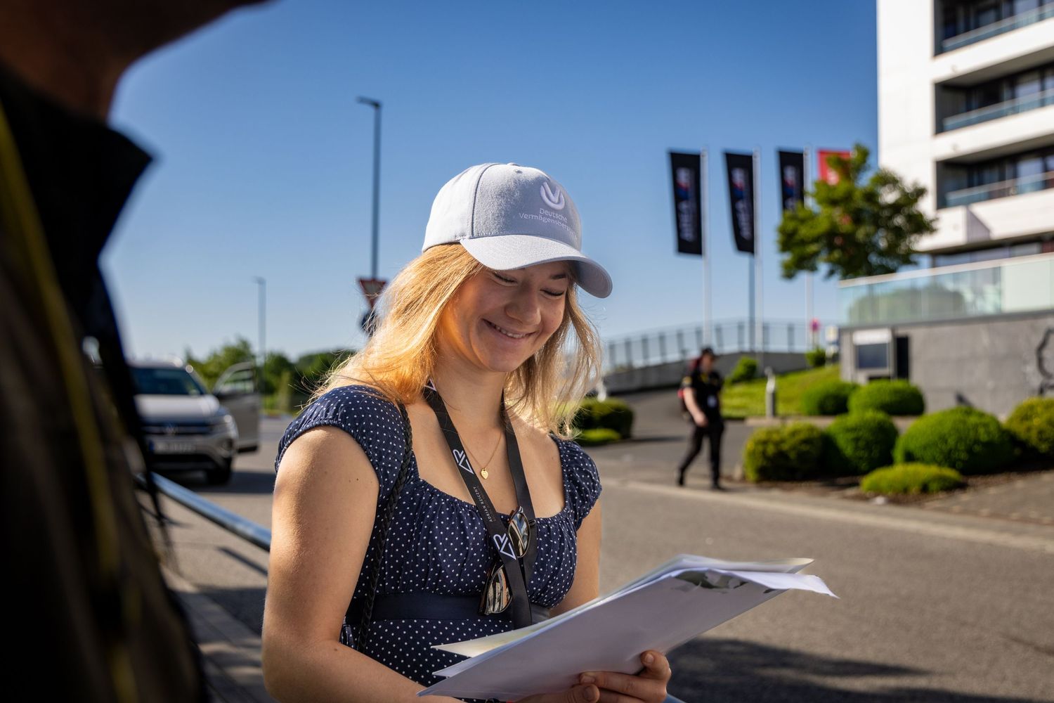Smiling blonde woman in white cap and polka dot dress reviewing documents outdoors on a sunny day.
