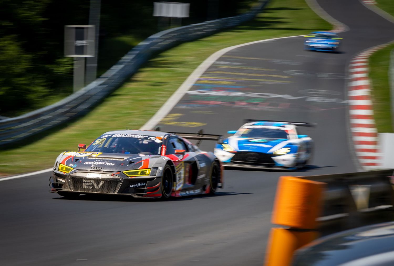 Racing cars compete on a winding track surrounded by lush green trees during a GT motorsport event.