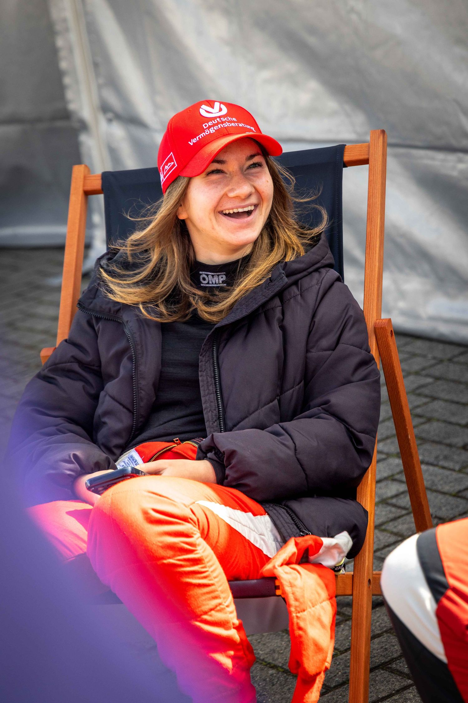 Woman in red cap and black jacket laughing while relaxing in a deck chair outdoors.