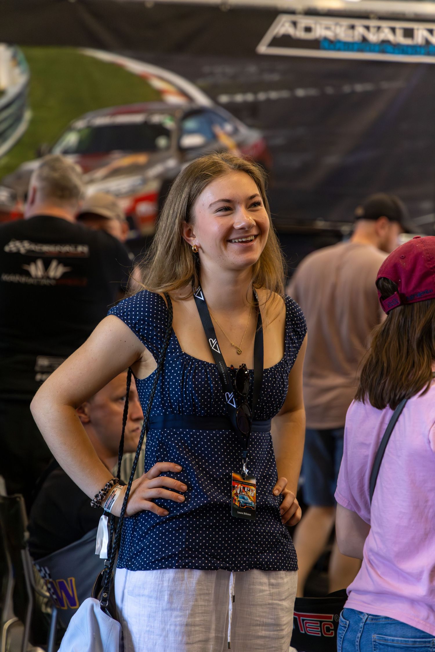 Smiling young woman in polka dot top with lanyard at an Adrenalin motorsport event, standing confidently with hand on hip.