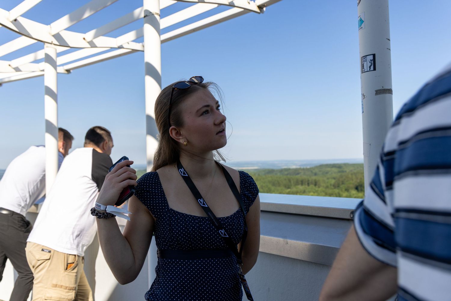 Woman in polka dot dress holding drink on rooftop terrace with scenic landscape view on sunny day.
