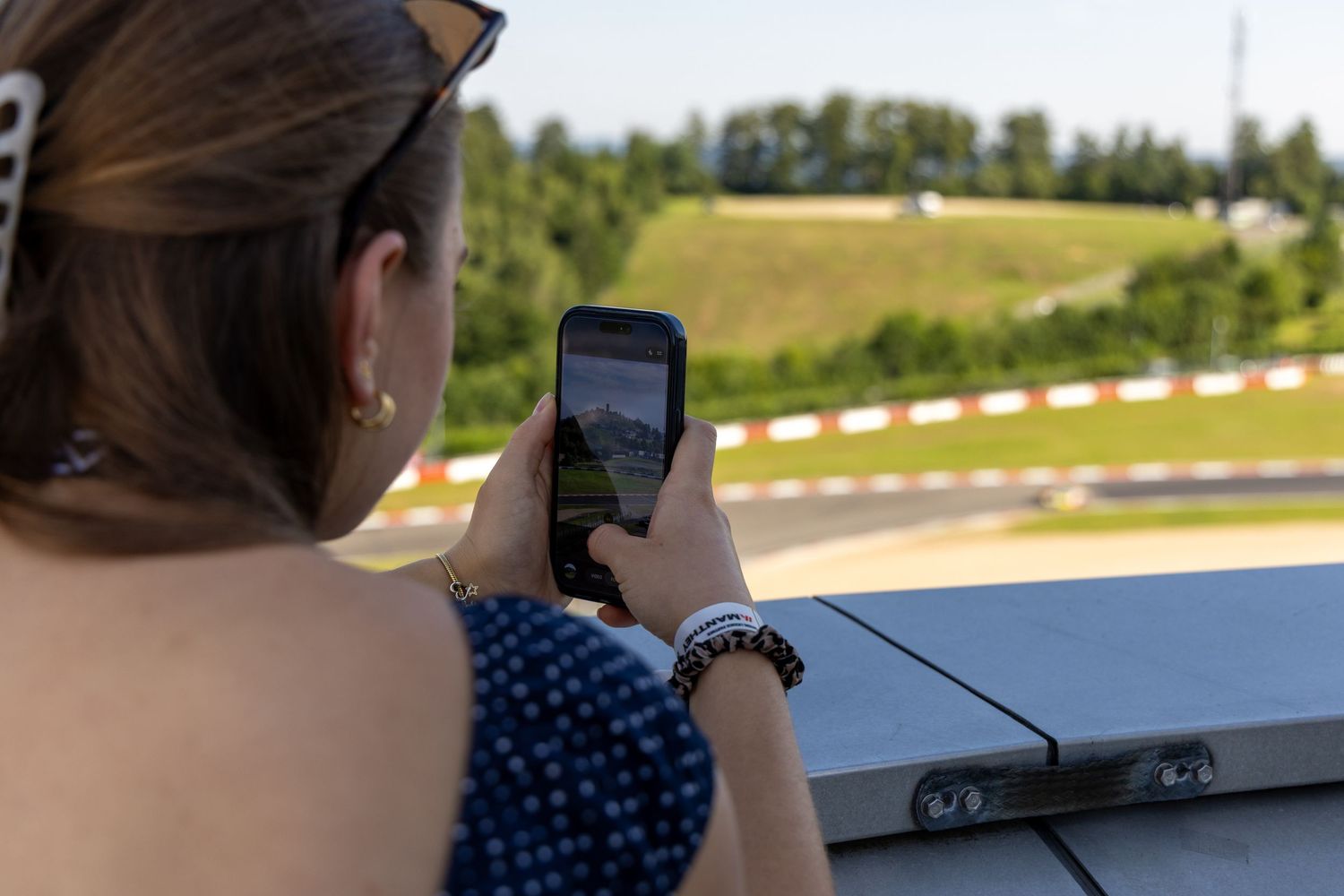 Woman photographing a racing circuit track with her smartphone from an elevated grandstand viewpoint on a sunny day.