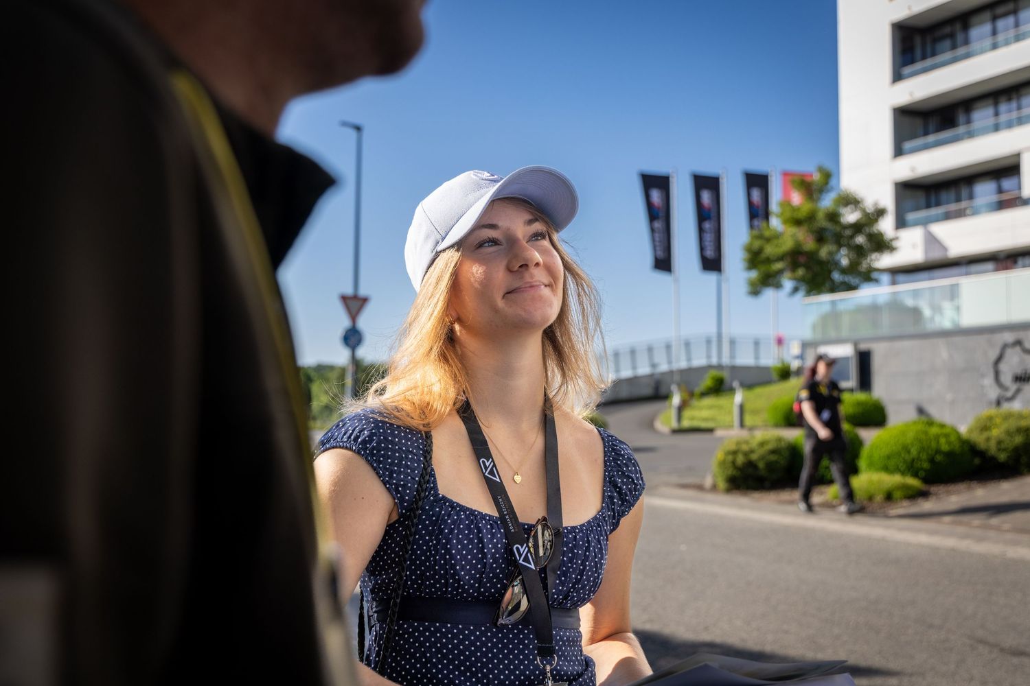 Blonde woman in white cap and blue polka dot dress smiling outdoors on a sunny day near a modern building.